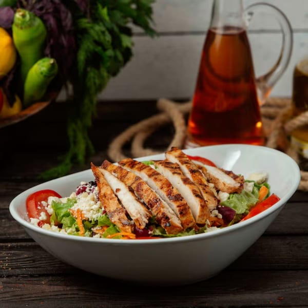 Grilled chicken salad with lettuce, tomatoes, cheese, and peppers in a white bowl.