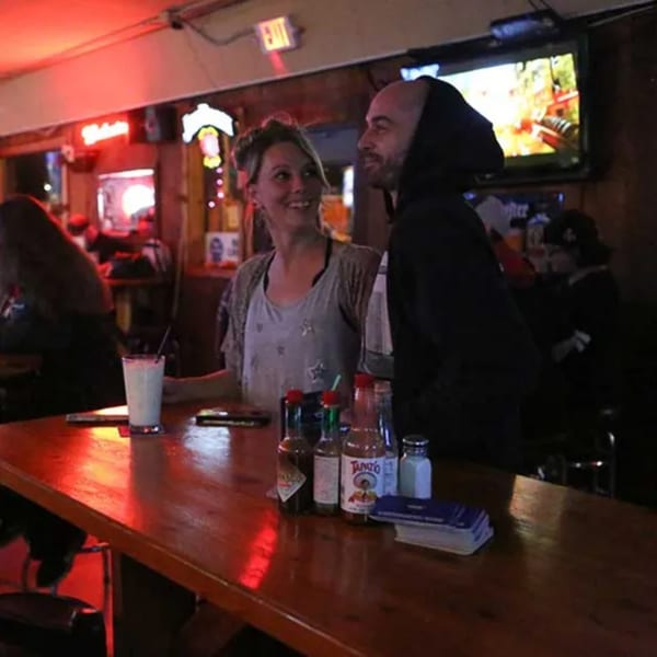 A couple smiles at each other while sitting at a bar with drinks and hot sauce bottles.