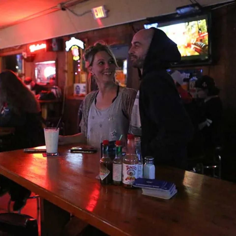 A couple smiles at each other while sitting at a bar with drinks and hot sauce bottles.